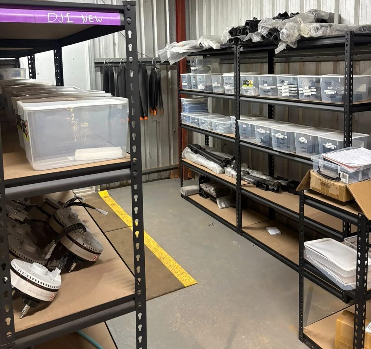 The AGX parts store: racking with labelled tubs of T50 and T100 spare parts, propeller blades hanging on the wall and wrapped arms on the top shelf